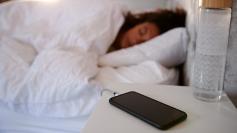 Smartphone lädt auf einem Nachttisch neben einem Glas Wasser, während im Hintergrund eine Person in einem Bett schläft. Die Umgebung ist ein Schlafzimmer mit weißen Bettdecken.