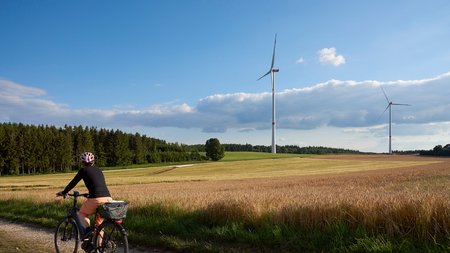 Eine Person fährt mit einem Fahrrad auf einem Feldweg durch eine ländliche Szene. Im Hintergrund stehen zwei Windkraftanlagen neben Feldern und Wäldern unter einem blauen, leicht bewölkten Himmel.