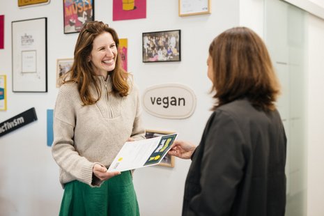 Eine Frau überreicht einer anderen Frau ein Zertifikat. Sie lächeln in einem Büro mit bunten Postern an der Wand. Das Wort "vegan" ist sichtbar.
