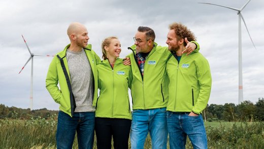 Vier Menschen stehen vor einer Windenergie-Anlage und schauen sich lachend an. Sie tragen hellgrüne Jacken mit dem Logo von Green Planet Energy.
