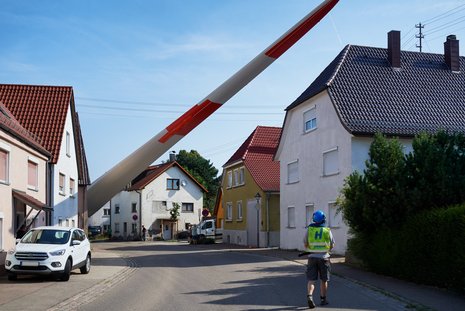 Ein Windradflügel wird durch eine enge Dorfstraße transportiert. Im Vordergrund ein Arbeiter, der sich den Flügel ansieht.