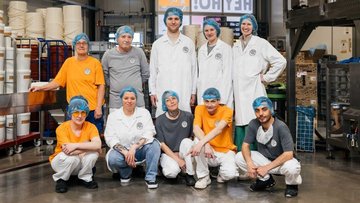 Ein Team von neun Personen trägt Arbeitskleidung und Haarnetze. Sie posieren lächelnd in einer Produktionsumgebung mit Regalen und Maschinen im Hintergrund.