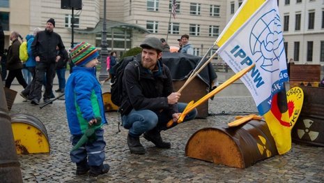 Mann hält Flaggen von "IPPNW" und Anti-Atomkraft auf öffentlicher Versammlung; Kind mit Hut daneben. Im Hintergrund sehen Passanten auf gepflastertem Platz vor historischen Gebäuden zu. Text auf Flagge: "IPPNW Internationale Ärzte für die Verhütung des Atomkrieges, Ärzte in sozialer Verantwortung e.V."