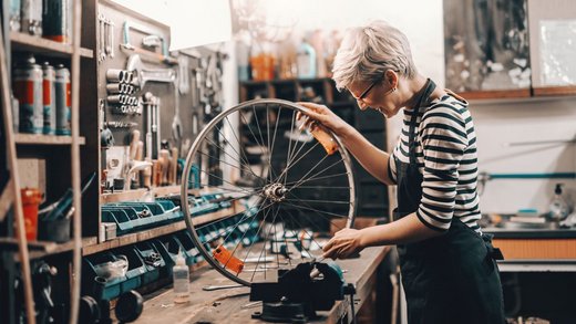 Eine Frau mit weißen blonden Haaren und in gestreiftem Shirt mit schwarzer Schürze steht an einer Werkbank und hält einen Fahrradrahmen in der Hand.