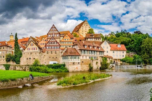 Fachwerkhäuser stehen an einer Flussböschung in einer historischen Stadt. Im Vordergrund sitzen zwei Menschen auf einer Wiese, während Wolken den Himmel bedecken.