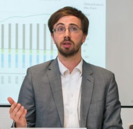 Ein Mann mit Brille in weißem Hemd und grauem Sakko steht vor einer an die Wand projizierten Grafik und hält einen Vortrag 