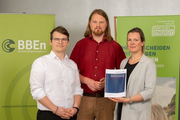 Zwei Männer und eine Frau, die ein DINA4 großes Papier sichtbar hochhält, stehen nebeneinander und blicken in die Kamera