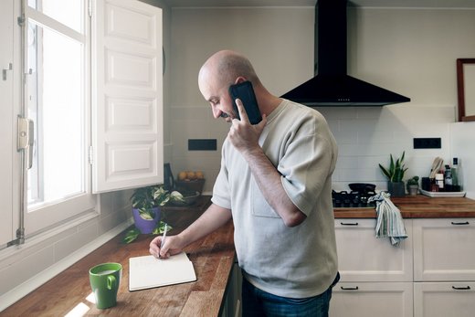 Ein Mann steht in einer Küche und hält sich mit einer Hand ein Telefon ans Ohr, während er mit der anderen Hand Notizen auf einem Notizblock öffnet.