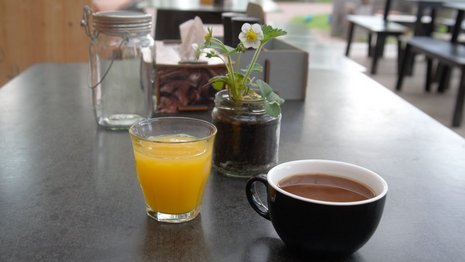 Ein Glas Orangensaft und eine Tasse Kaffee stehen auf einem Tisch in einem Café. Daneben sind ein Einmachglas mit einer Pflanze und Servietten zu sehen. Im Hintergrund Sitzgelegenheiten.