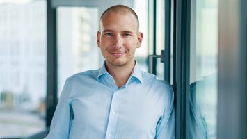Ein Mann in einem hellblauen Hemd lehnt lächelnd an einer Glaswand in einem hellen Bürogebäude. Sein Spiegelbild ist in der Glaswand sichtbar.