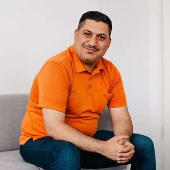 Ein Mann dunklen kurzen Haaren sitz auf einem hellgrauen Sofa und lächelt in die Kamera. Er hat seine Arme auf die Oberschenkel gestützt und die Hände zusammengelegt. Er trägt ein orangenes Polo-Shirt und eine dunkelblaue Jeans.