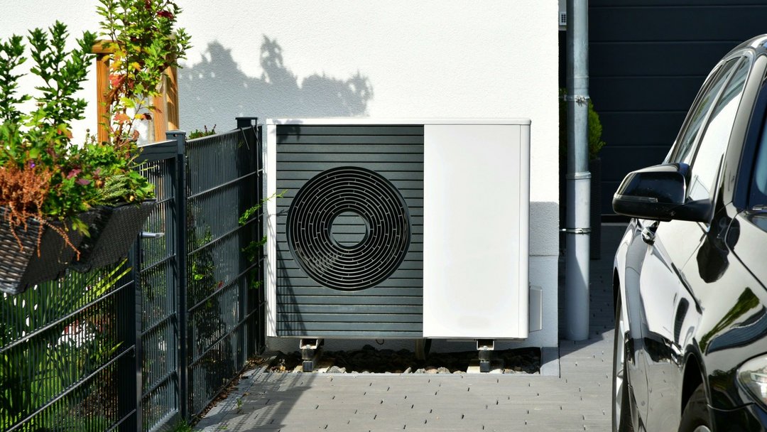 Wärmepumpe steht in der Einfahrt eines Hauses. Daneben ist ein ein Zaun mit Pflanzen und ein Teil eines schwarzen Autos zu sehen.