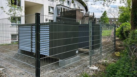 Zwei große Wärmepumpeneinheiten stehen in einem umzäunten Bereich neben einem Wohngebäude. Im Hintergrund sind Balkone, Grünflächen und Bäume zu sehen.