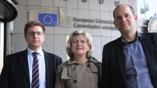 Drei elegant gekleidete Personen blicken in die Kamera. Im Hintergrund das Gebäude der Europäischen Kommission.