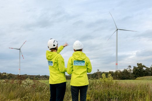 Zwei Mitarbeitende von Green Planet Energy mit weißem Baustellen-Helmen in hellgrünen Jacken mit dunkelgrünem Logo stehen auf einem Feld vor zwei Windkraftanlagen. Die linke Person zeigt auf eins der Windräder