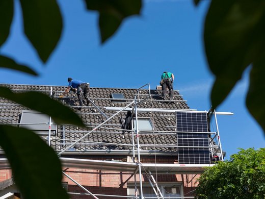 Zwei Personen bauen eine Solaranlage auf ein schräges Hausdach. An der Seite des Hauses ist ein Gerüst angebracht.