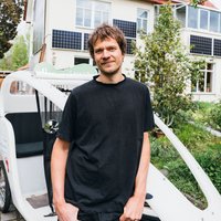 Eine Person steht neben einem weißen Elektrofahrzeug. Im Hintergrund befindet sich ein Haus mit mehreren Solarpaneelen an der Fassade. Es ist eine grüne, bewachsene Umgebung.