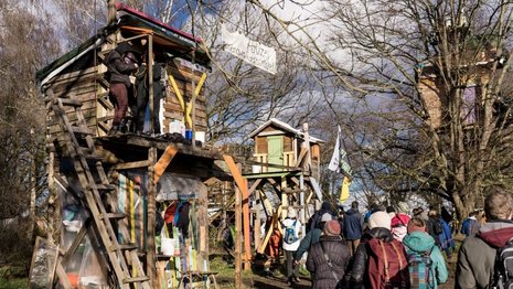 Holzhäuser stehen auf Stelzen, umgeben von Menschen in Outdoor-Kleidung. Ein Transparent mit der Aufschrift "Polizei Arschkarte" ist sichtbar. Die Szene spielt in einer bewaldeten, sonnigen Umgebung.