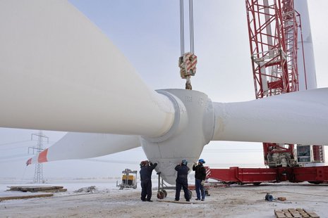 Die drei Flügel der Windkraftanlage werden von einem roten Kran angehoben, während drei Arbeiter den Vorgang überwachen.