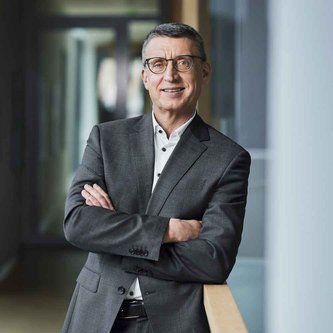 Ein Mann mit dunkelgrauen kurzen Haaren und eckiger schwarzer Brille steht mit verschränkten Armen an ein Geländer gelehnt. Er trägt einen grauen Anzug und lächelt in die Kamera.