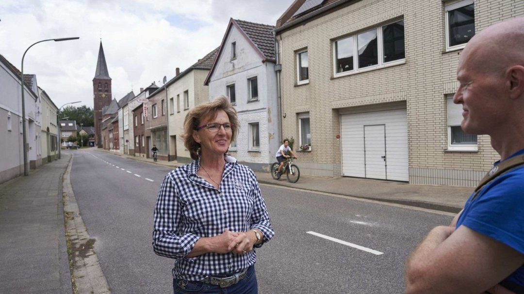 Eine Frau spricht mit einem Mann in einer Wohnstraße mit Kirchenhintergrund. Ein Radfahrer passiert die Szene. Ziegelhäuser und bewölkter Himmel umgeben die Personen. Keine sichtbare Beschilderung oder Text.