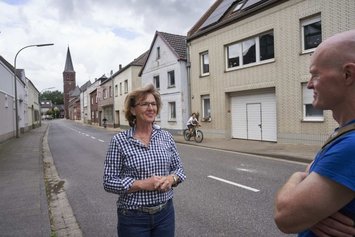 Eine Frau spricht mit einem Mann in einer Wohnstraße mit Kirchenhintergrund. Ein Radfahrer passiert die Szene. Ziegelhäuser und bewölkter Himmel umgeben die Personen. Keine sichtbare Beschilderung oder Text.
