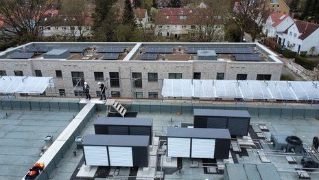 Auf dem Flachdach des vorderen Gebäudes stehen mehrere große Wärmepumpen. Dahinter liegt ein weiteres Gebäude, dessen Dach mit Photovoltaikmodulen in Reihen belegt ist. Im Hintergrund sind Wohnhäuser und Bäume zu erkennen.