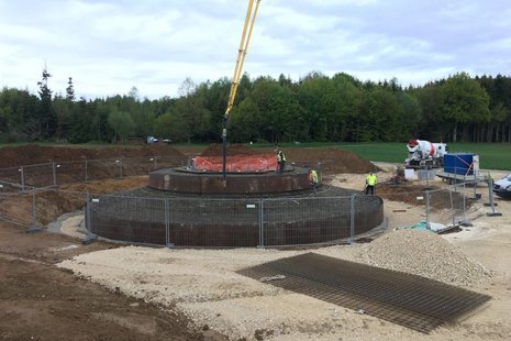 Rundes Erdloch mit einem umgebenden Zaun und 3 Arbeitern und einem Baufahrzeug.