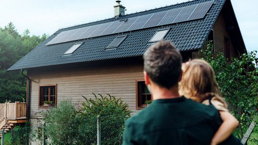 Ein Mann in dunkelgrünem Pullover trägt ein blondes Kind auf dem Arm und steht mit dem Rücken zur Kamera. Vor ihnen steht ein Holzhaus mit einer PV-Anlage auf dem Dach, das sie sich anschauen.