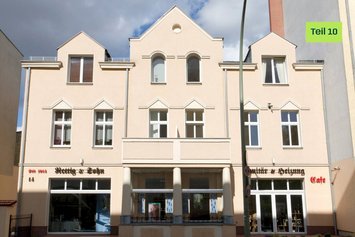 Ein helles Gebäude mit drei Giebeln, davor einzelne grüne Straßenlaterne. Im gelben Kreis steht „Blogserie Teil 10“. Oben rechts: „Green Planet Energy“. Unten sind Ladenschilder zu sehen.