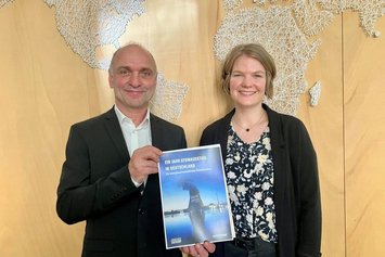 Zwei Personen halten ein Dokument mit dem Titel „Ein Jahr Atomausstieg in Deutschland“ vor einer hölzernen Weltkarte in einem Büro oder Konferenzraum. Das Bild strahlt Professionalität aus.