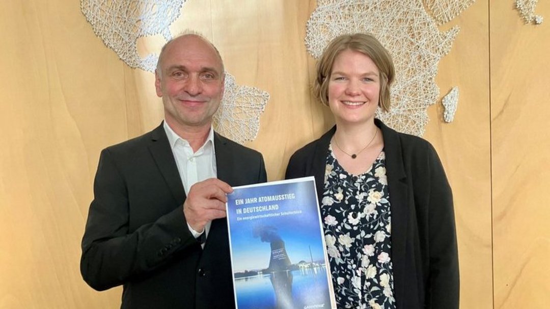 Zwei Personen halten ein Dokument mit dem Titel „Ein Jahr Atomausstieg in Deutschland“ vor einer hölzernen Weltkarte in einem Büro oder Konferenzraum. Das Bild strahlt Professionalität aus.