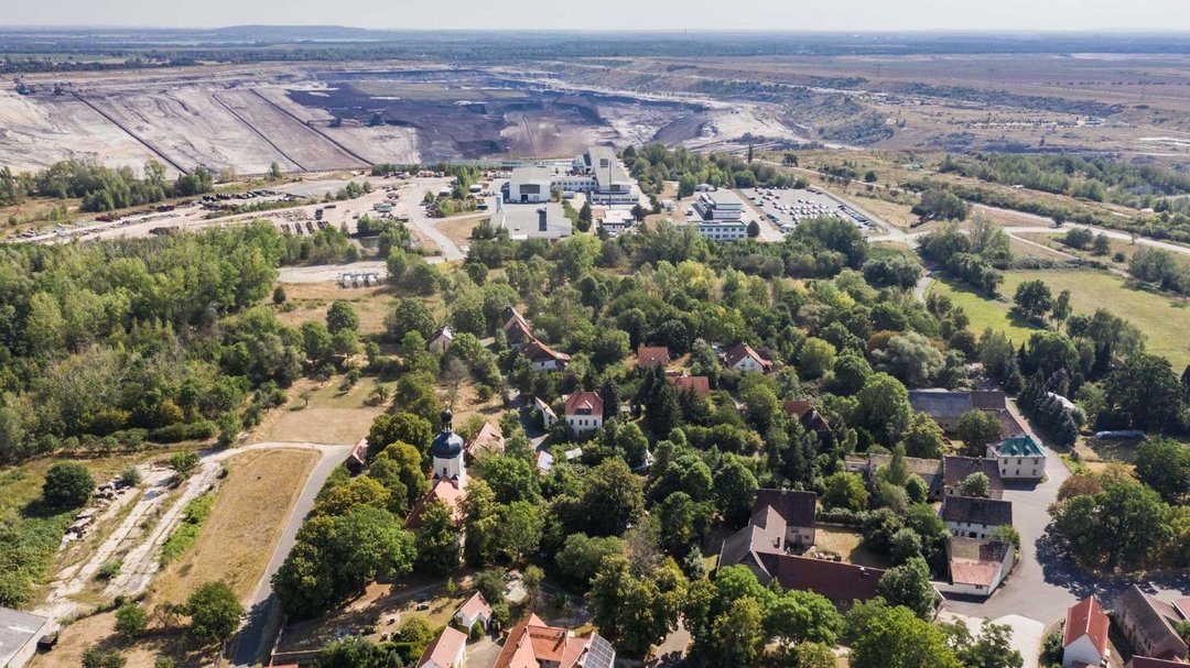 Luftaufnahme von einem Dorf bei Sonnenschein. Im Hintergrund ist ein kahles Kohlebaugebiet zu sehen.