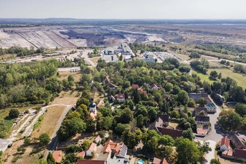 Luftaufnahme von einem Dorf bei Sonnenschein. Im Hintergrund ist ein kahles Kohlebaugebiet zu sehen.