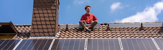 Mann sitzt auf einem Hausdach oberhalb einer Photovoltaikanlage