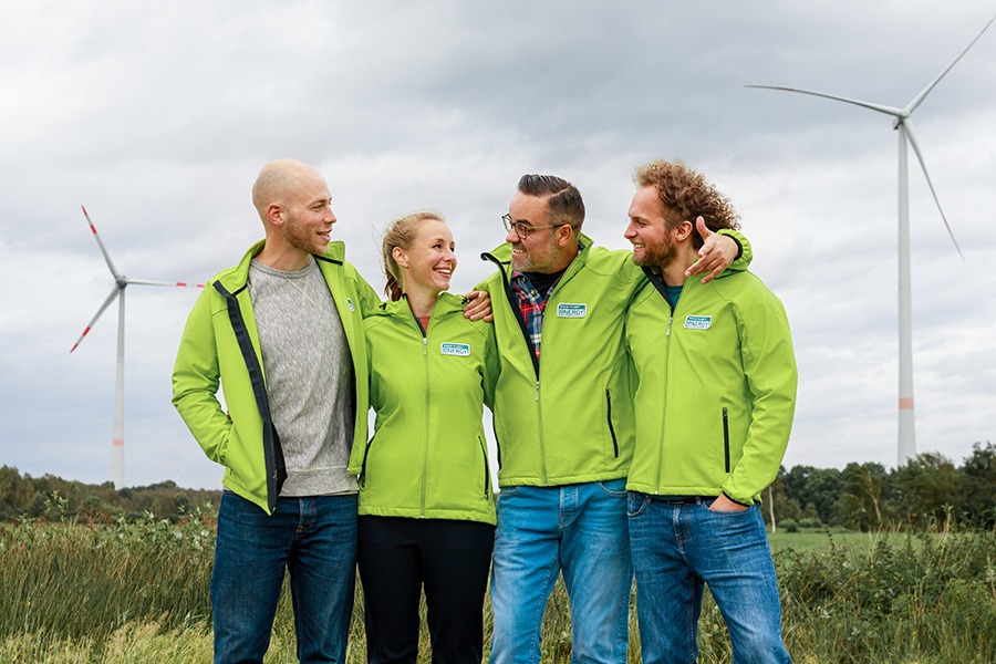 Vier Personen mit einer Grüne Jacke von Green Planet Energy unterhalten sich. Im Hintergrund ein Feld und zwei Windräder.